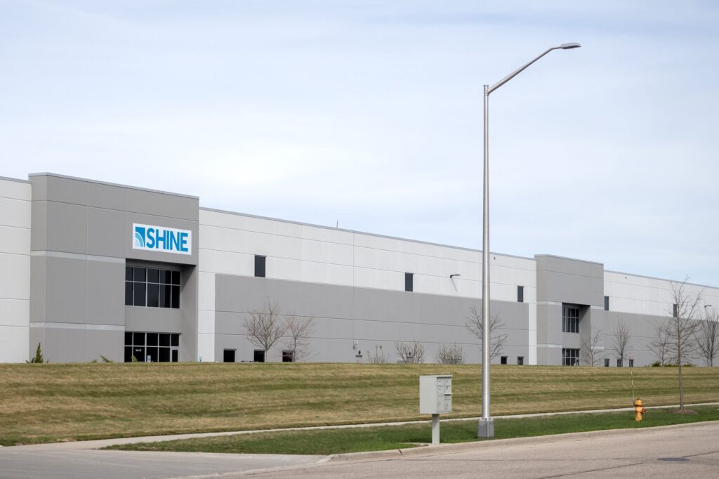 A large industrial building with a blue roof labeled ISHINE, with several parked cars in front and an open field in the foreground.
