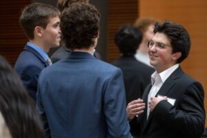 Three men in suits stand in a group, engaged in conversation at an indoor event.