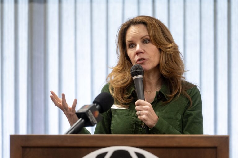 A woman speaks into a microphone at a podium, gesturing with one hand. A microphone is positioned on the podium in front of her. Vertical blinds cover the window behind her.
