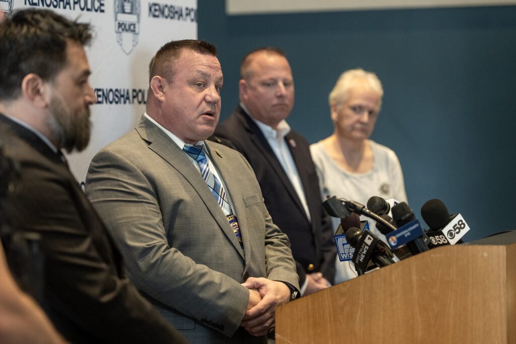 Black and white portrait of an older man with short hair, wearing dark-rimmed glasses and a serious expression, facing the camera.