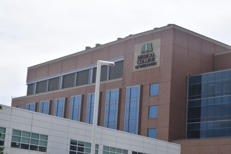 A man in a dark suit with a light pink shirt and tie stands smiling in front of a blurred background with green and white tones.
