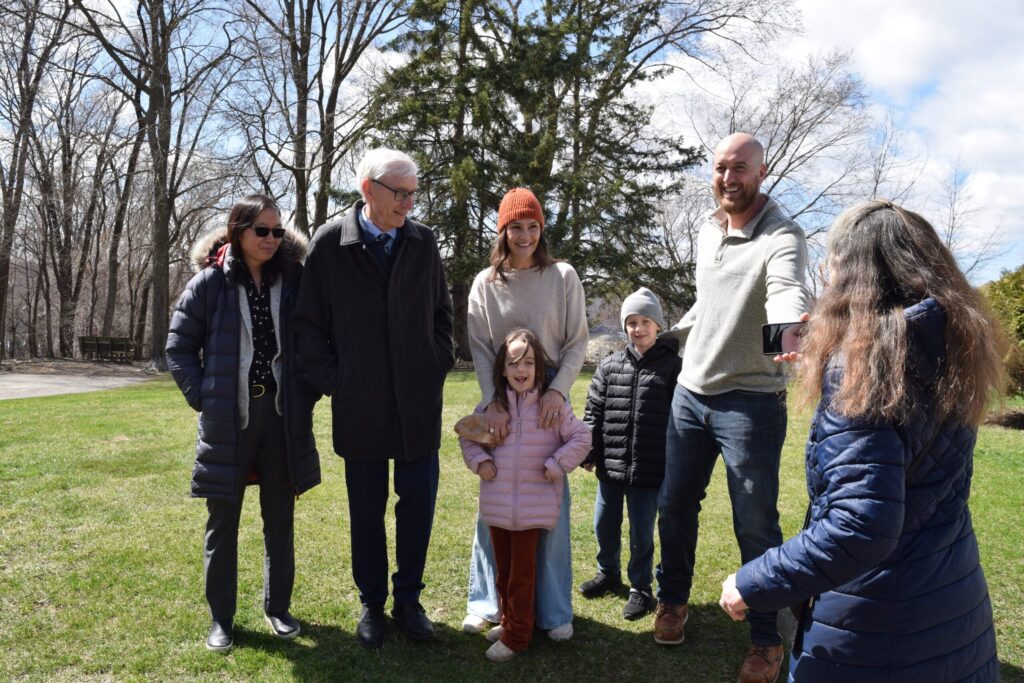 Four people stand outside near a white building, talking on a sunny day with trees and blue sky in the background.