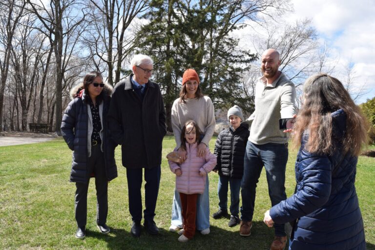 Four people stand outside near a white building, talking on a sunny day with trees and blue sky in the background.