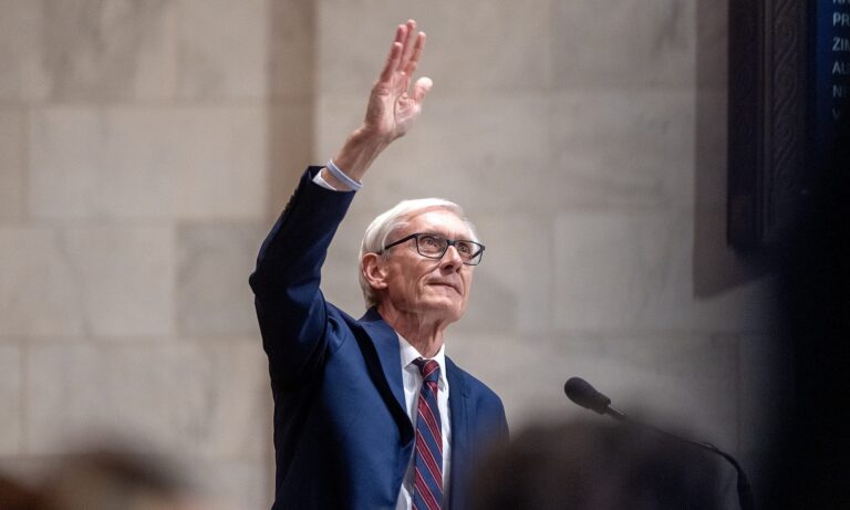 An older man in a suit and striped tie raises his right hand while standing at a podium in front of a stone wall.
