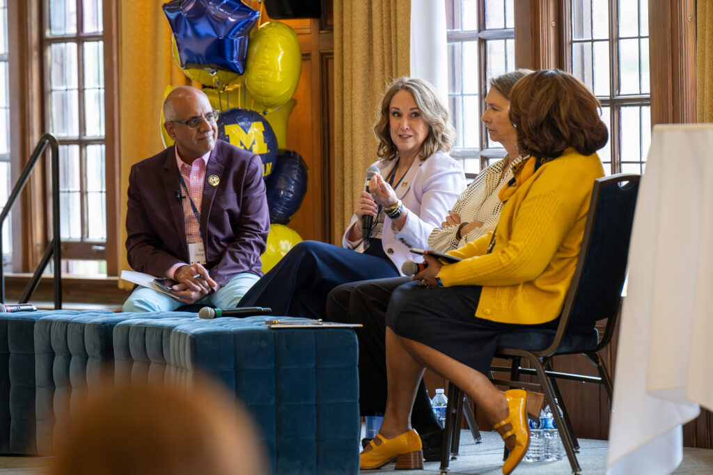 Four people sit in chairs on a stage and speak to a group of people