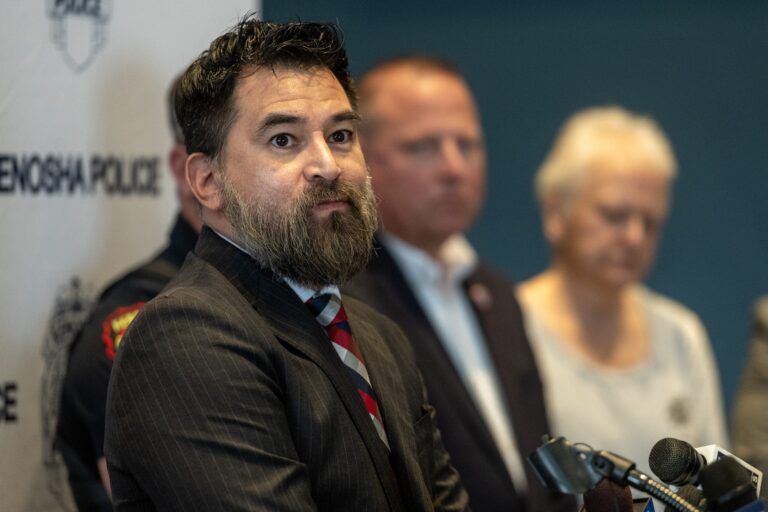 A man in a suit stands at a podium during a press conference, with three other people and a Kenosha Police banner in the background.