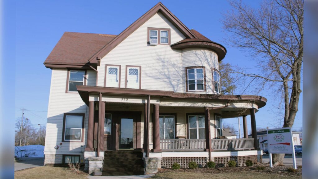 A hallway with dark wood trim features a door to the outside, a window, and two side doors. There are small tables with plants and a lamp, and an EXIT sign above the door.