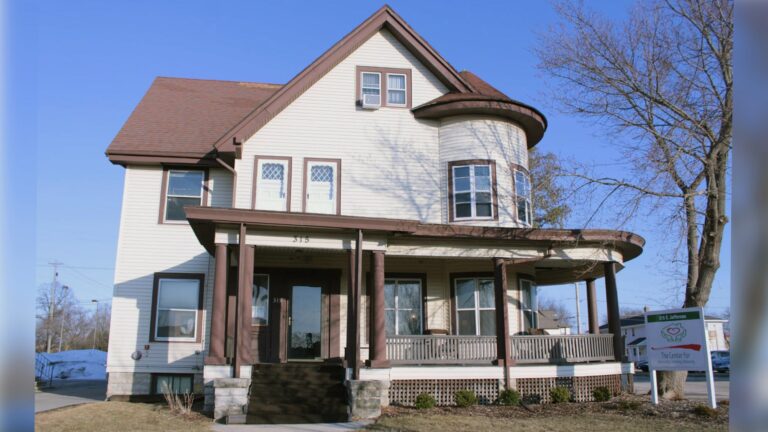 A hallway with dark wood trim features a door to the outside, a window, and two side doors. There are small tables with plants and a lamp, and an EXIT sign above the door.