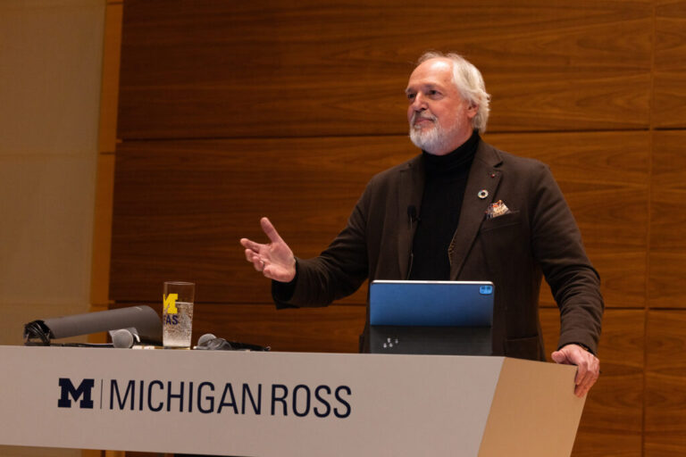 A man gestures with one hand while speaking behind a lectern that says Michigan Ross