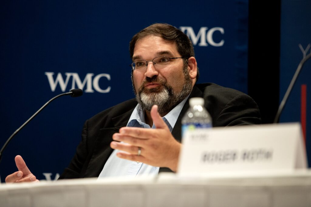 A man with glasses and a beard speaks at a panel discussion, gesturing with his hand. A microphone and water bottle are in front of him, with a blurred name card in the foreground.