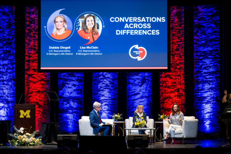 A man and two women sit in chairs on a stage