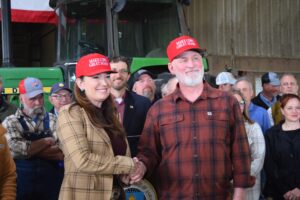 People wearing red hats sit at tables in a barn-like setting, participating in a meeting near a green John Deere tractor and a large pile of feed or hay.