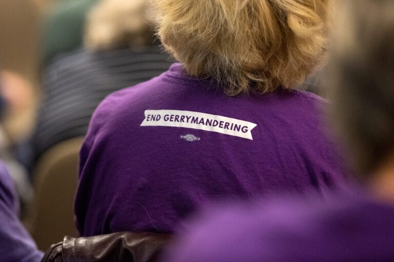 Four women stand side by side, smiling and waving in front of a blank projection screen in a conference room.