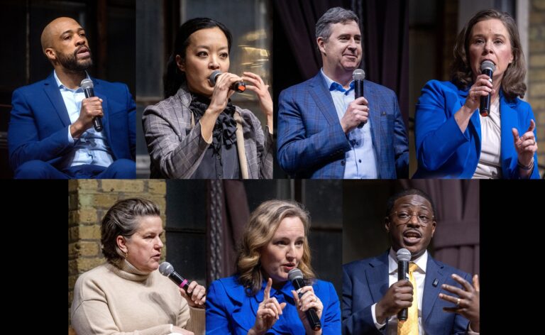 Seven people sit and speak into microphones during what appears to be a panel discussion or forum, wearing business attire and seated against a brick wall backdrop.