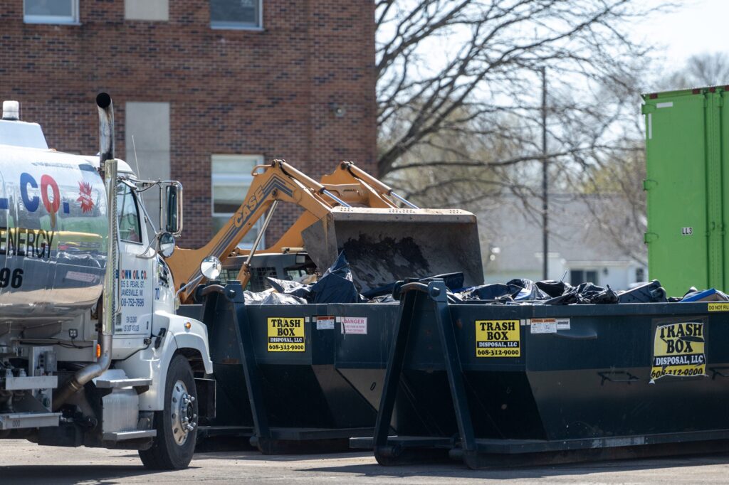 A black pickup truck drives through a flooded residential street with houses and trees in the background under a cloudy sky.