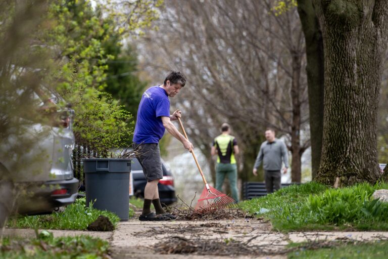 A person uses a chainsaw to cut fallen tree branches in a yard, surrounded by debris and broken limbs.