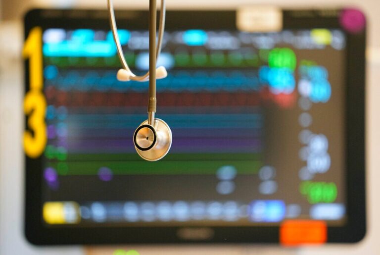 A stethoscope hangs in front of a medical monitor displaying patient vital signs with colorful graphs and numbers.