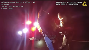 A sheriff’s deputy detains a person at night on a road, illuminated by police vehicle lights; body camera footage displays timestamp and police details.