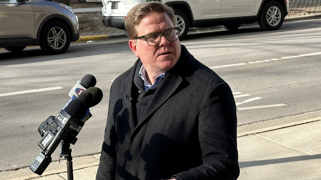 A man in a dark coat stands outdoors next to microphones from various news outlets, with parked cars and a street in the background.