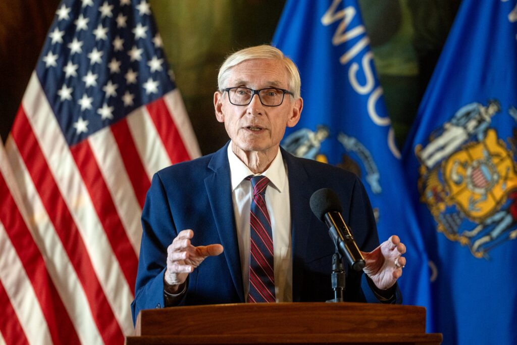 An older man in a suit and tie speaks at a podium with a microphone, with American and Wisconsin state flags in the background.