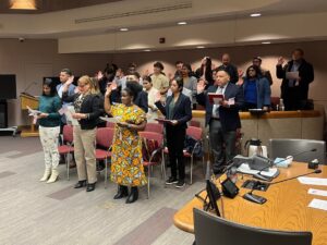Griselda Solis de Druckers Journey to US Citizenship and Love - Common Good News A group of eight adults stands indoors against a blue wall, with one woman holding a bouquet and official papers. Everyone is smiling at the camera.