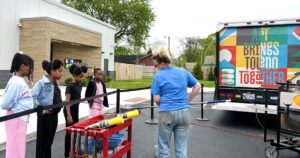 Kacey McCreery, in a blue t-shirt and red sunglasses, smiles in front of the “Baby Dragon”, the truck behind her reading “Art Brings Toledo Together”.