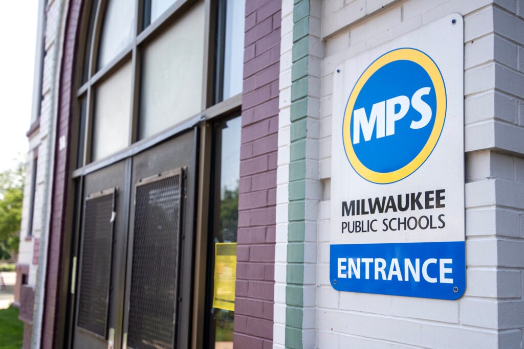 A sign reading Milwaukee Public Schools Entrance is mounted on a white brick wall next to a large windowed door.