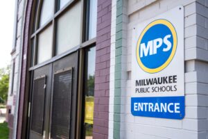 A sign reading Milwaukee Public Schools Entrance is mounted on a white brick wall next to a large windowed door.