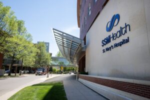 Exterior view of SSM Health St. Mary’s Hospital with a building sign, sidewalk, trees, and a skywalk connecting buildings.