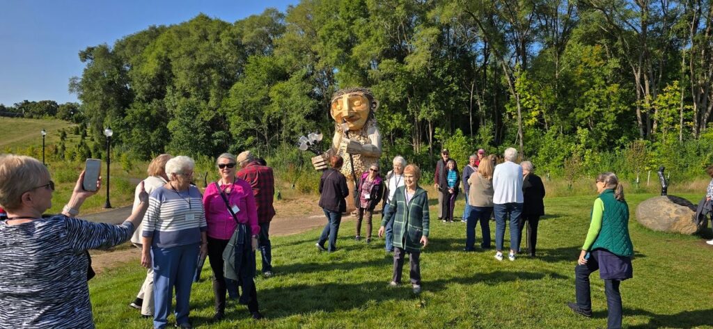 A man in a red jacket stands on a path with a large wooden troll sculpture and trees in the background.