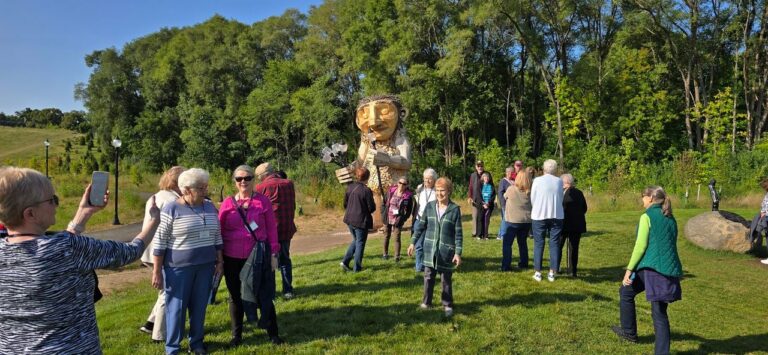 A man in a red jacket stands on a path with a large wooden troll sculpture and trees in the background.