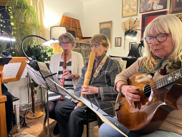 Three older women sit indoors reading sheet music, two playing recorders and one playing an acoustic guitar, surrounded by plants and artwork on the walls.