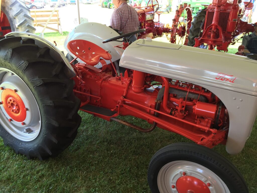 A vintage Ford tractor with red and gray paint is on display outdoors, surrounded by grass and other tractors.