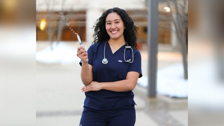 A woman in a white dress and gold graduation stole stands outdoors, smiling and holding a stethoscope.
