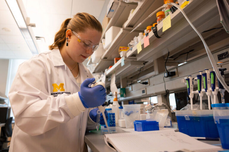 A student in a lab coat and goggles with the Block M on them pipes liquid into a beaker at a bench filled with equipment and notes at the Parent Lab in the Life Sciences institute.