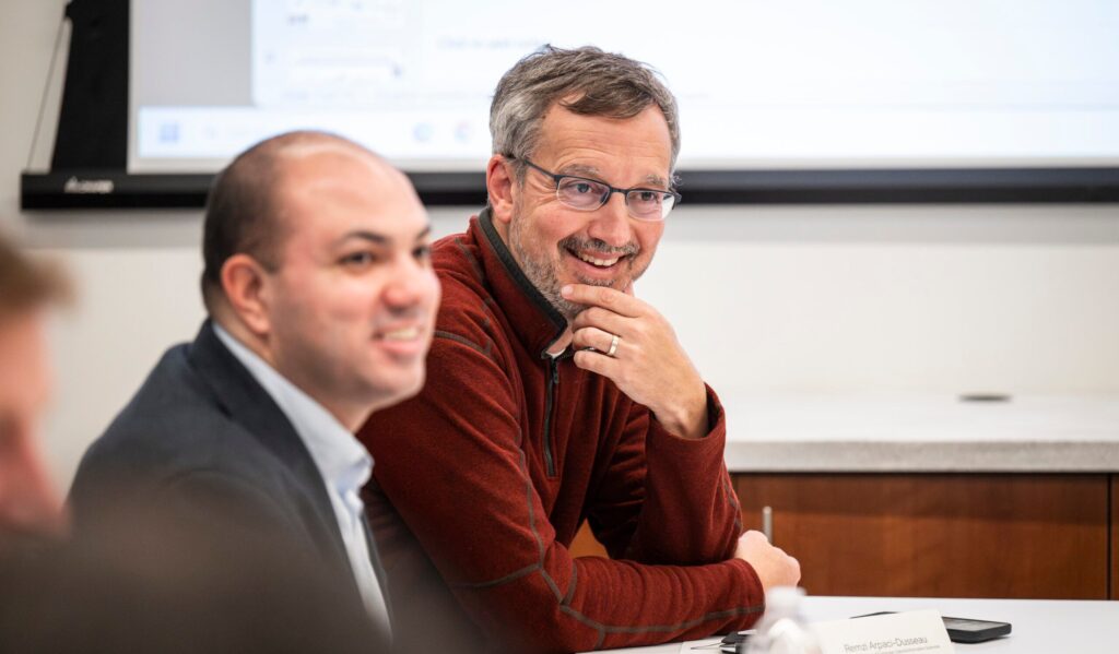 Two men sit at a conference table during a meeting, with one man smiling and the other looking ahead; paperwork and a water bottle are on the table.