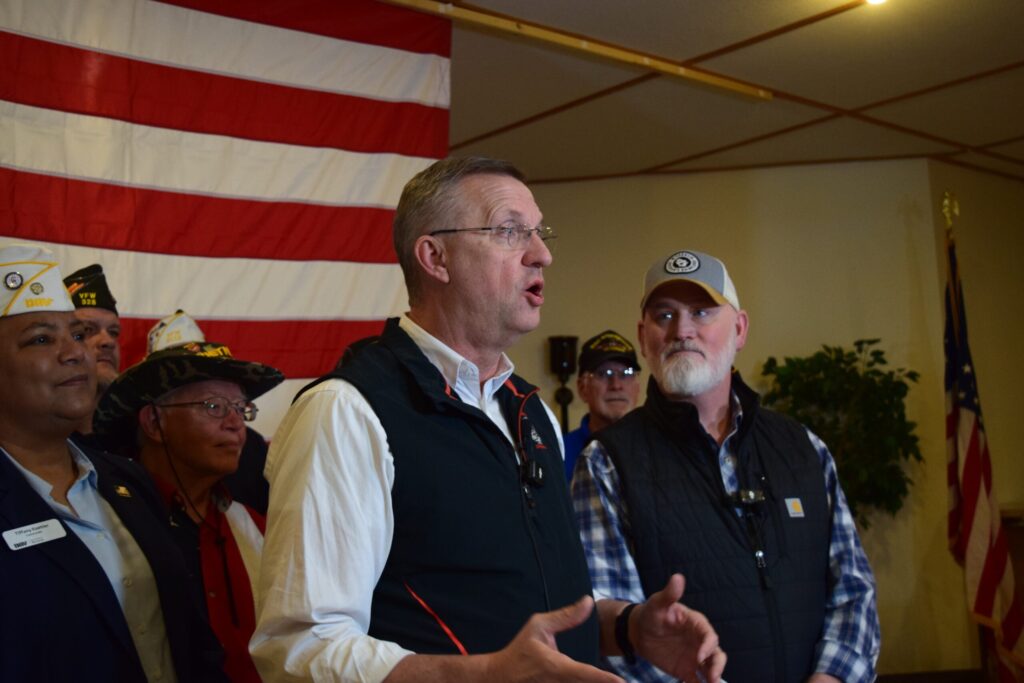 A group of people, some in military uniforms, stand indoors in front of a large American flag, with one man speaking in the foreground.