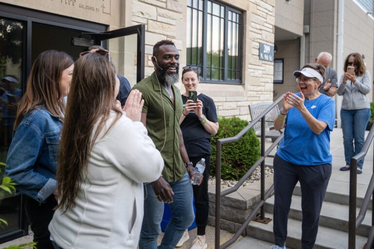 A man steps outside a building as a group of people stand around him, clapping and smiling; some are taking photos with their phones.