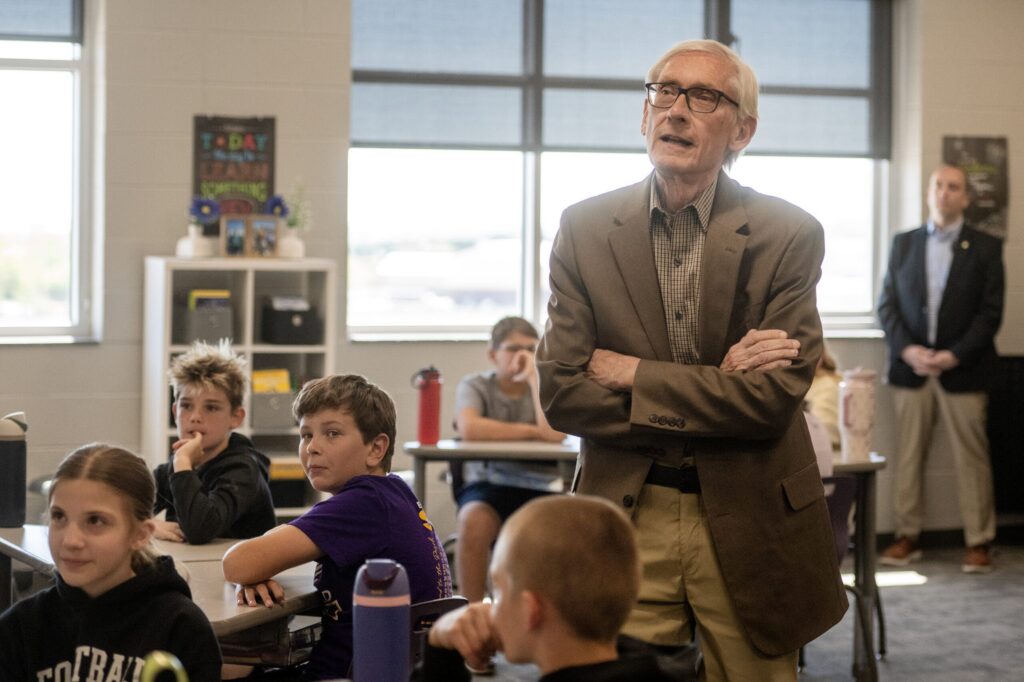 An older man in a suit jacket stands speaking in a classroom, while students sit at desks and listen. An adult stands in the background near the window.