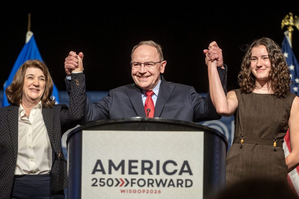 An older man in a suit and striped tie speaks at a podium labeled AMERICA with a microphone, against a background with Anderson and dates visible.