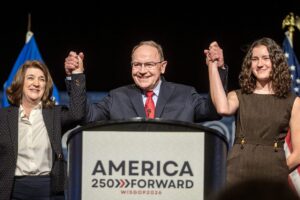 An older man in a suit and striped tie speaks at a podium labeled AMERICA with a microphone, against a background with Anderson and dates visible.