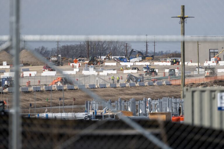 Construction site with workers, vehicles, and machinery operating; fencing and materials visible in the foreground.