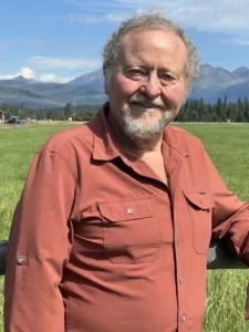 Smiling older man with gray hair and beard in a rust shirt, standing in a grassy field with mountains in the background.