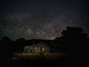 Night scene with a weathered wooden cabin beneath a star-filled sky and the Milky Way, surrounded by trees at dusk.