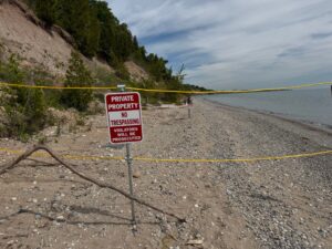 Controversy Erupts Over Private Property Signs at Lions Den Gorge - Common Good News A red and white Private Property No Trespassing sign is posted on a sandy beach, with yellow rope marking the boundary. Trees and a steep shoreline are in the background.