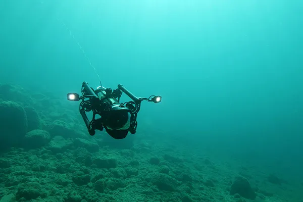 An underwater drone with lights navigates above a rocky seabed in clear blue water.