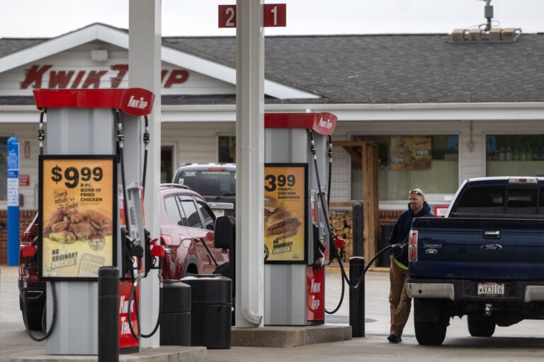A man stands outside his pickup truck at a gas station with red pumps, near a Kwik Trip store; promotional food signs are visible on the pumps.