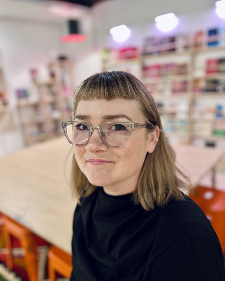 A person with shoulder-length light brown hair, glasses, and a black shirt sits in a room with shelves and a wooden table in the background.