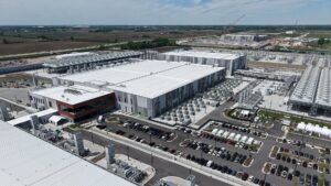 Aerial view of a large industrial facility with several white-roofed buildings, parking lots filled with cars, and farmland in the background.