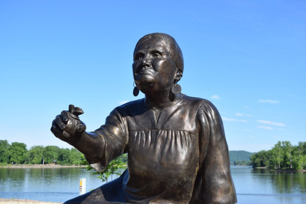 Bronze statue of a woman kneeling with one arm extended, situated outdoors by a river with trees and a lamppost in the background.